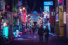 people walking down an alley way at night with neon signs on the buildings in the background