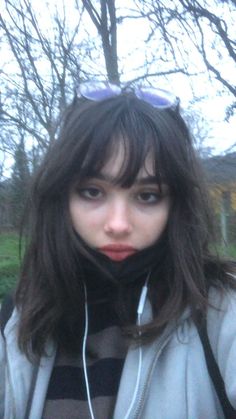 a young woman with earphones on standing in front of some trees and grass, looking at the camera