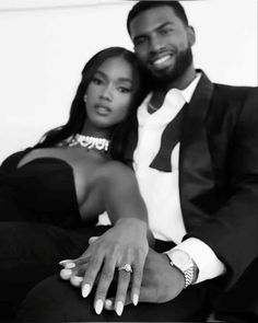 a man and woman sitting next to each other in front of a white wall with their arms around each other