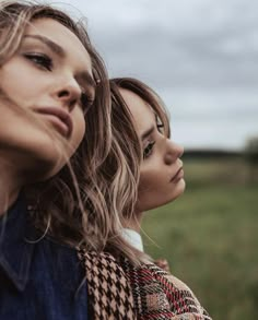 two beautiful women standing next to each other in a field with grass and clouds behind them