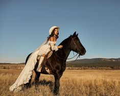 a woman dressed in white riding on the back of a brown horse across a dry grass field