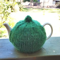 a green knitted tea pot cover sitting on top of a wooden table next to a tree