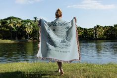 a woman is holding up a towel in front of a body of water with palm trees behind her