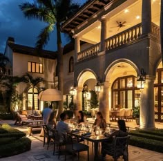 people are sitting at a table outside in front of a large house with palm trees