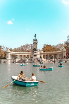 two people in a row boat on the water near a fountain with statues behind them