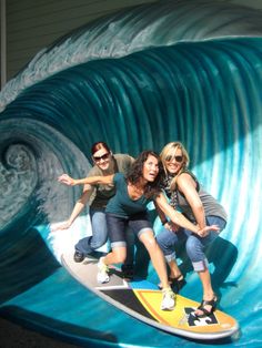 three women are posing on a surfboard in front of a giant wave sculpture at the beach