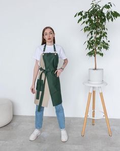 a woman standing in front of a potted plant wearing an apron and white t - shirt