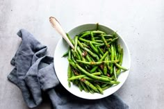 a white bowl filled with green beans on top of a blue cloth next to a wooden spoon