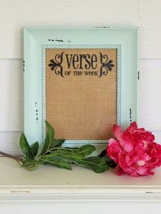 a pink flower sitting on top of a shelf next to a framed sign that says verse of the week