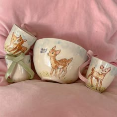 three cups with deer designs on them sitting on a pink bed sheet next to a pillow