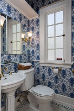 a white toilet sitting next to a bathroom sink under a window with blue wallpaper