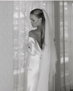 a woman in a wedding dress standing next to a window with her veil pulled back