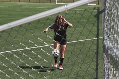 a woman in black shirt kicking a soccer ball on green field with fence and grass