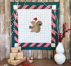 a quilted wall hanging with a squirrel holding a christmas present in front of it