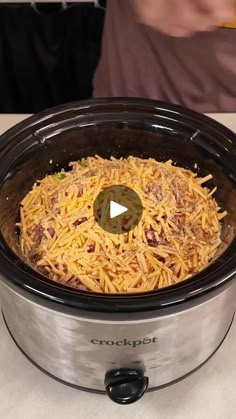 a crock pot filled with shredded cheese on top of a white countertop next to a person