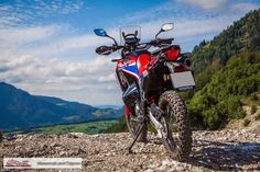 a red, white and blue motorcycle parked on the side of a rocky road with mountains in the background