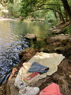 a blanket, book and shoes are sitting on the edge of a river bank next to some rocks