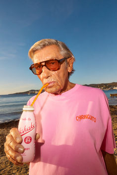 an old man with sunglasses and a pink shirt is drinking from a can on the beach