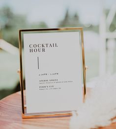 a cocktail hour sign sitting on top of a wooden table next to a white feather