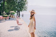 a woman in a hat and sunglasses standing on the edge of a pier by the water