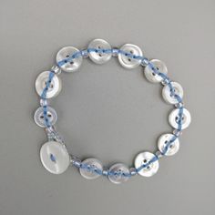 a white and blue beaded bracelet with buttons on the clasp is shown against a gray background