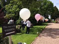 some people are standing in the grass with balloons on their heads and one is holding a sign that says this way to the wedding