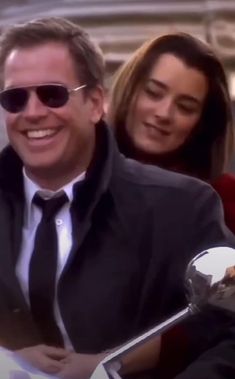 a man in a suit and tie smiles as he sits on a motorcycle with two women behind him