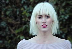 a woman with blonde hair and pink lipstick looks at the camera while standing in front of some bushes