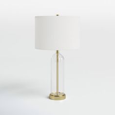 a glass table lamp with a gold base and white linen shade on the top, against a plain background