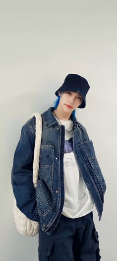 a young man with blue hair wearing a denim jacket and hat, standing against a white wall