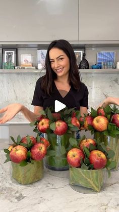 a woman sitting at a table with several vases filled with apples and leaves on it