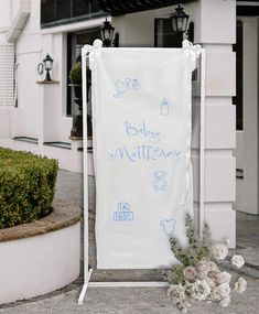 a white sign sitting on the side of a road next to a bush and flowers