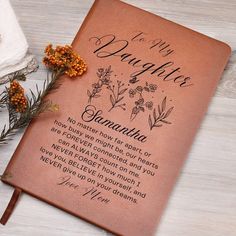 a brown leather book with writing on it and some flowers next to it, sitting on top of a white wooden table