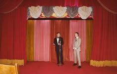 two men in tuxedos standing on stage with red curtains and drapes behind them