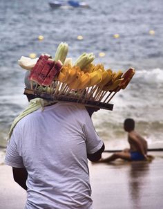 a man walking on the beach carrying a basket filled with bananas and other fruit items