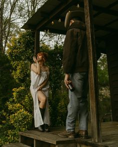 a woman in a white dress is standing on the porch next to a man with a cowboy hat