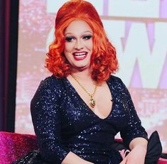 a woman with red hair sitting on a pink chair and smiling at the camera while wearing a black sequined dress