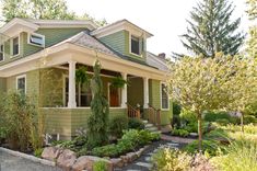 a green house with white trim on the front porch and covered in greenery, shrubs and trees