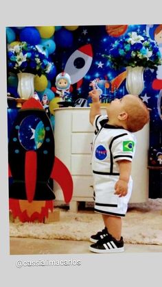 a baby boy standing in front of a rocket ship themed wall with balloons and decorations