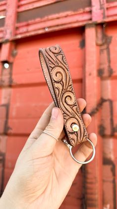 a hand holding a leather keychain in front of a red brick wall with an intricate design on it