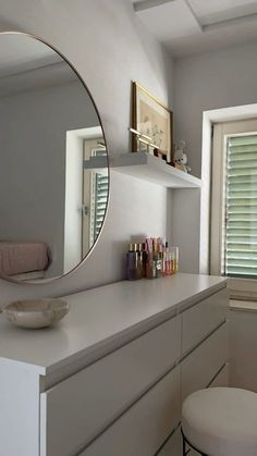 a white bathroom with a round mirror above the vanity and stool next to it, in front of a window