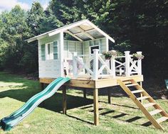 a small white house with a slide in the grass next to some trees and bushes