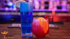 two colorful drinks sitting next to each other on top of a wooden table in front of a bar