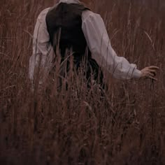 a person standing in tall grass with their hands out to the side, wearing a white shirt and black vest