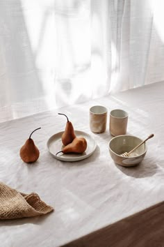 three pears and two bowls on a white tablecloth with curtains in the background