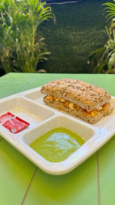 a sandwich on a white plate with green soup in front of it and some plants