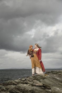 two people standing on rocks near the ocean under a cloudy sky with an eagle perched on their arm