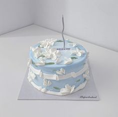 a blue cake with white flowers on it sitting on top of a card board in front of a wall