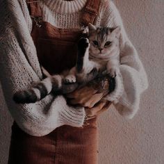 a woman holding a cat in her arms while wearing overalls and a sweater over her shoulders