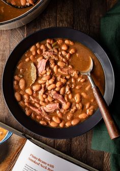 a black bowl filled with beans and ham next to a book on a wooden table